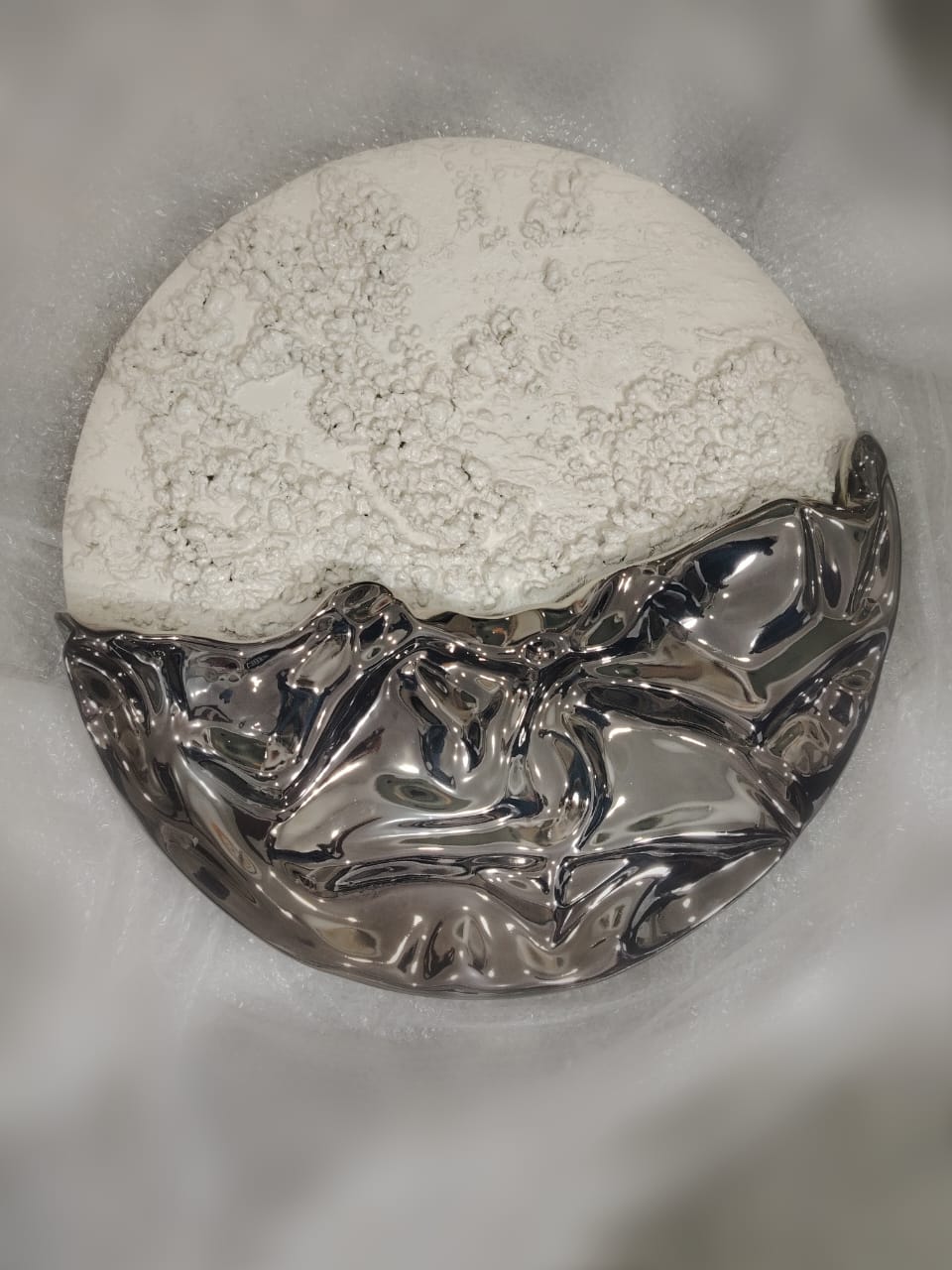A round wall sculpture: textured white plaster above a rippling wave of polished silver